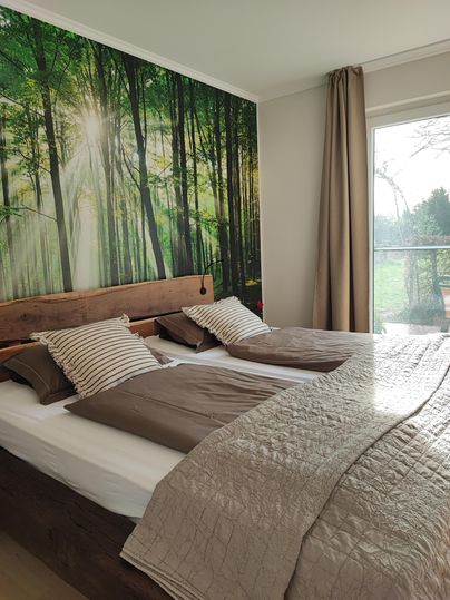 A cozy bedroom with a double bed and a wooden panel wall. The forest background and natural light create a relaxed atmosphere.