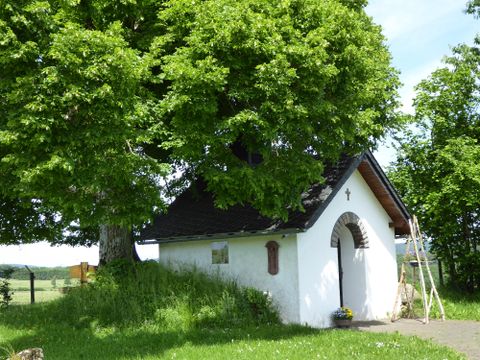 A small, white chapel stands under a large tree. Surrounded by green grass and an open landscape.