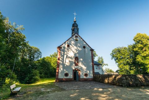 Une petite église avec un clocher et une croix au sommet. Entourée d'arbres verts et d'un ciel bleu clair.