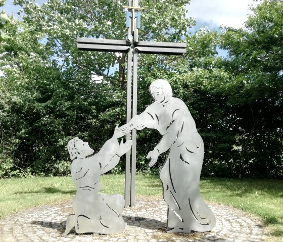 Sculpture en m&eacute;tal avec une croix et deux personnages, l'un agenouill&eacute;, l'autre debout, sur un cercle pav&eacute; dans un jardin verdoyant.