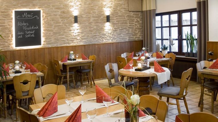 A cozy restaurant with wooden chairs and tables. The tables are decorated with white tablecloths and red napkins.