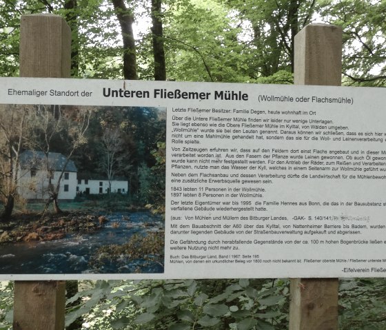 Een bord in het bos geeft informatie over de Untere Flie&szlig;emer M&uuml;hle. Het toont een foto van de molen en bevat historische details over het gebruik en de geschiedenis ervan., &copy; TI Bitburger Land