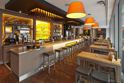 Ein modernes Gebäude mit Glasfronten und mehreren Geschäftseinheiten. Draußen stehen Tische und Pflanzen, die eine einladende Atmosphäre schaffen.