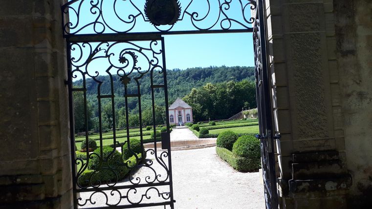 Une belle vue à travers un portail en fer forgé sur un jardin bien entretenu et un bâtiment charmant en arrière-plan. Des arbres verts et un ciel bleu complètent le paysage.