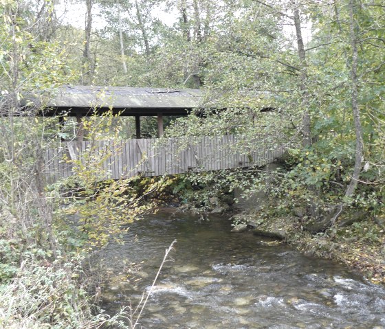 Een overdekte houten brug steekt een kleine rivier over in een bosrijke omgeving. Herfstbomen omringen het tafereel., &copy; Felsenland S&uuml;deifel Tourismus GmbH, Christian Calonec-Rauchfuss