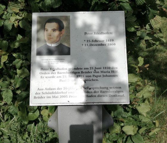Plaque comm&eacute;morative pour Peter Friedhofen avec photo et texte, entour&eacute;e de feuilles vertes., &copy; TI Bitburger Land