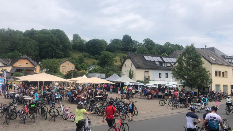 Een druk plein met veel mensen en fietsen. Op de achtergrond zijn huizen en bomen te zien, evenals parasols.
