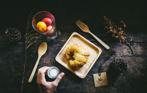 A rustic table with a wooden top, on which a pastry and a glass bowl with fresh fruit are placed. One hand holds a small glass while a spoon lies next to it.