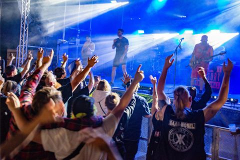 Een grote menigte juicht tijdens een live concert. Het podium is verlicht en de artiesten optreden met energie.