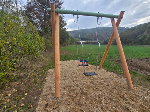 Une balançoire se trouve sur un sol sableux pavé dans un parc tranquille. En arrière-plan, on peut voir des arbres et des collines douces.