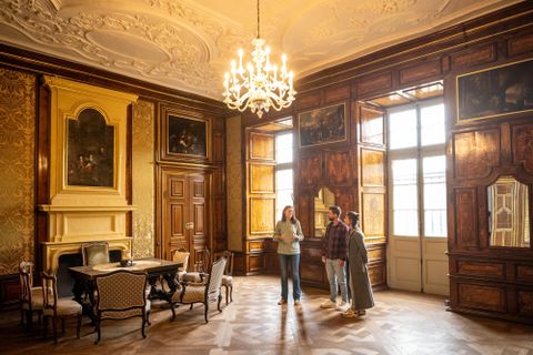 Ein elegantes Zimmer mit Holzwänden und einem großen Kronleuchter. Drei Personen stehen am Tisch und betrachten die Einrichtung.