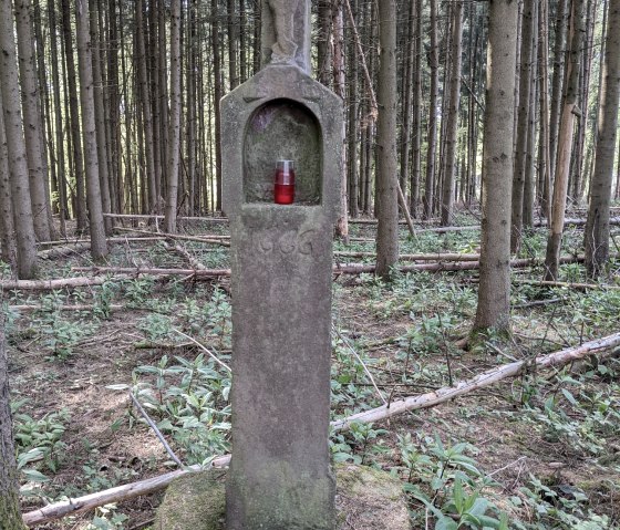 Une croix de chemin en pierre avec une statue du Christ et une lampe fun&eacute;raire rouge se trouve dans une for&ecirc;t dense., &copy; A. Girards