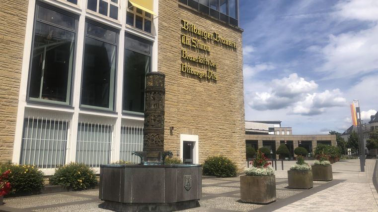 Un bâtiment moderne avec une grande façade en verre. Devant le bâtiment se trouve une fontaine ronde, entourée de bacs à plantes.