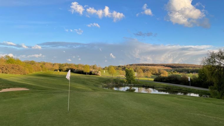 Eine malerische Golfplatzlandschaft mit grünem Rasen und einem kleinen Teich. Der Himmel ist blau mit einigen Wolken.