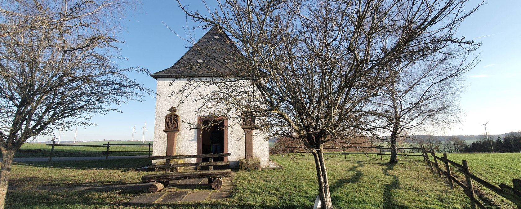Une petite chapelle sur une prairie verte, entour&eacute;e d'arbres d&eacute;nud&eacute;s. En arri&egrave;re-plan, on aper&ccedil;oit des &eacute;oliennes., &copy; TI Bitburger Land