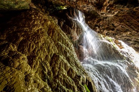 A waterfall cascades over a rocky cliff in a dark cave. The light reflects on the water, creating a mysterious atmosphere.