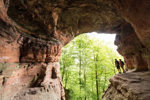 An impressive cave with red rocks and a view of a green forest. Two hikers stand at the cave's entrance, enjoying the scenery.