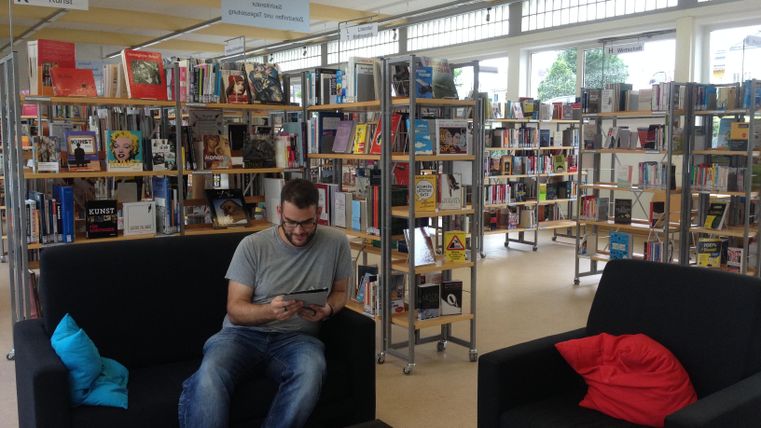 A man is sitting on a black sofa in a library. In the background, there are bookshelves filled with many books.
