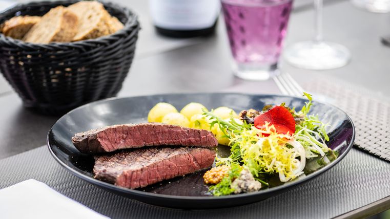 A beautiful plate with juicy steak, side dishes of potatoes and fresh salad. In the background, bread and a beverage can be seen.