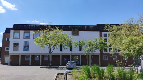 Een modern gebouw met een heldere façade en grote ramen. Voor het gebouw staan enkele bomen en het is een zonnige dag.