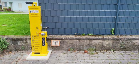 Een gele fietspomp staat tegen een muur. De vloer bestaat uit klinkers en er is groen gras op de achtergrond zichtbaar.