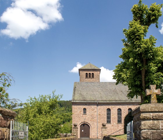Die Kirche St. Maximin in Kyllburg, umgeben von B&auml;umen, unter einem klaren blauen Himmel. Im Vordergrund sind steinerne Kreuze zu sehen., &copy; TI Bitburger Land