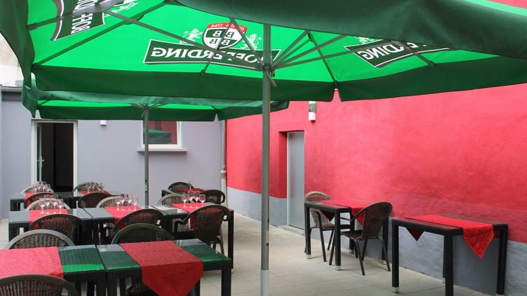 A cozy outdoor area with tables and chairs under green umbrellas. The walls are painted red, and the atmosphere is inviting.