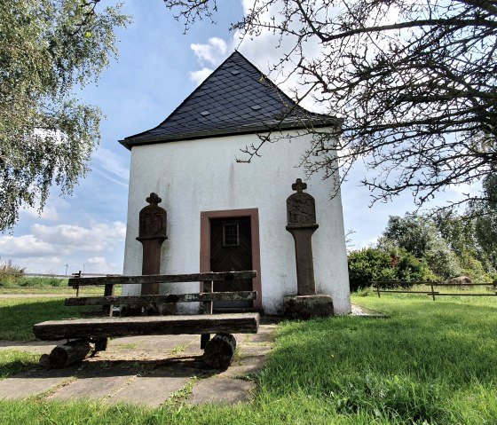 Wei&szlig;e Kapelle mit Schieferdach, flankiert von zwei Steins&auml;ulen. Eine Holzbank steht davor, umgeben von gr&uuml;ner Wiese und B&auml;umen., &copy; Tourist-Information Bitburger Land