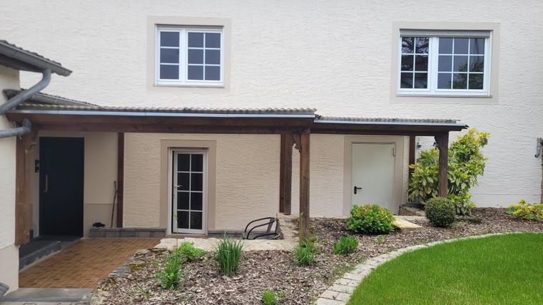 A modern house with a well-maintained green area. The entrance is surrounded by plants and a wooden veranda.