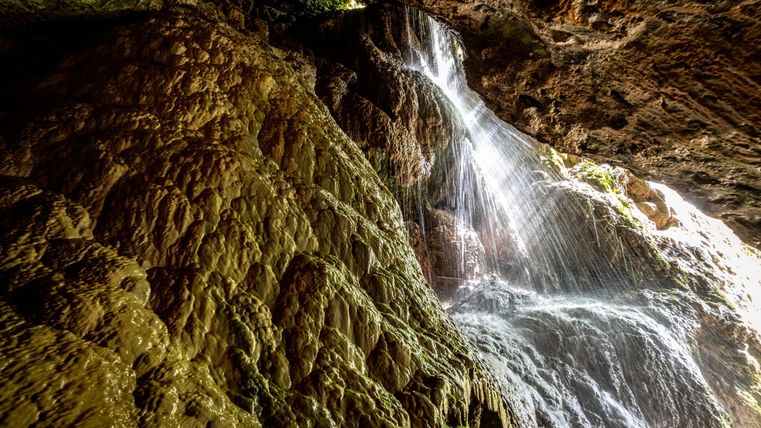 A waterfall cascades over a rocky cliff in a dark cave. The light reflects on the water, creating a mysterious atmosphere.