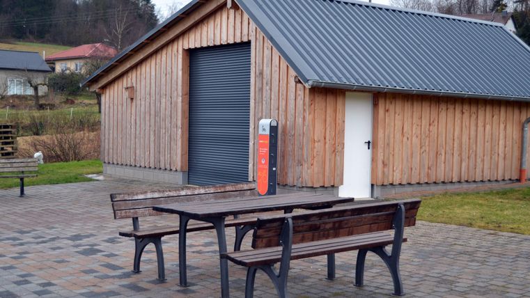 Een houten gebouw met een grote rolluik en een witte poort. Voor het gebouw staan twee houten tafels en banken op een betegeld gebied.