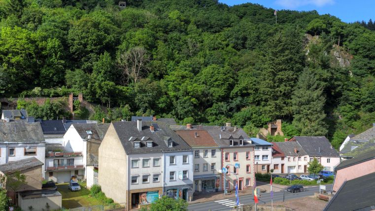 Een klein stadje met verschillende huizen ligt aan de voet van een groene heuvel. De blauwe lucht en de bomen geven de omgeving een rustige sfeer.