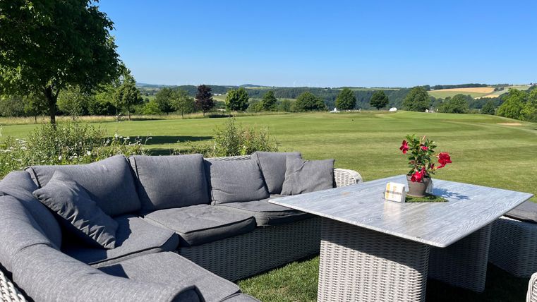 Ein gemütlicher Sitzbereich mit grauem Sofa und Tisch, umgeben von einer grünen Wiese. Im Hintergrund sind Bäume und ein klarer blauer Himmel zu sehen.