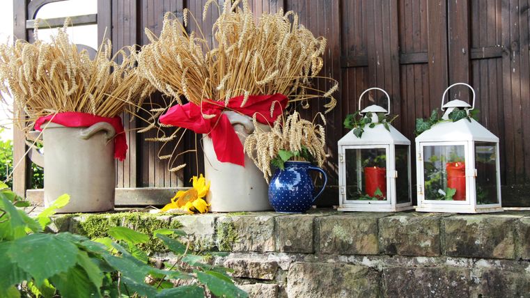 Two jugs with dried wheat and red ribbons stand next to two lanterns on a stone wall. In the background, a wooden wall is visible.