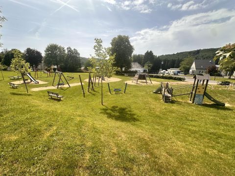 Ein Spielplatz mit verschiedenen Spielgeräten und viel grünem Gras. Im Hintergrund sind Bäume und einige benutzerfreundliche Sitzgelegenheiten zu sehen.