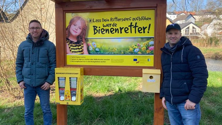 Twee mannen staan naast een informatieschild dat pleit voor de bescherming van bijen. Op de achtergrond zijn huizen en bomen zichtbaar.