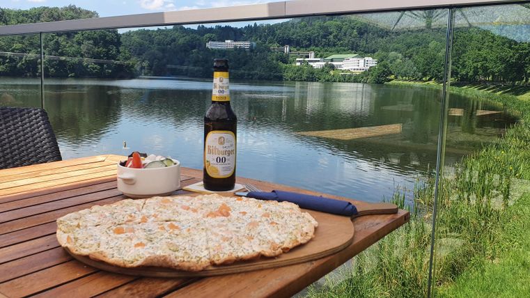Een tafel met een grote pizza, een fles bier en een kleine salade. Op de achtergrond is een schilderachtig meer en een groene landschap te zien.