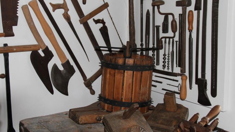 A collection of wooden tools is mounted on the wall. In the center stands a wooden barrel and a workbench with various tools.