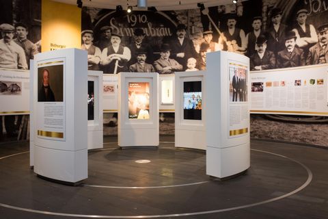 Eine Ausstellung mit mehreren weißen Displays, die historische Bilder präsentieren. Im Hintergrund sind historische Fotografien an der Wand zu sehen.