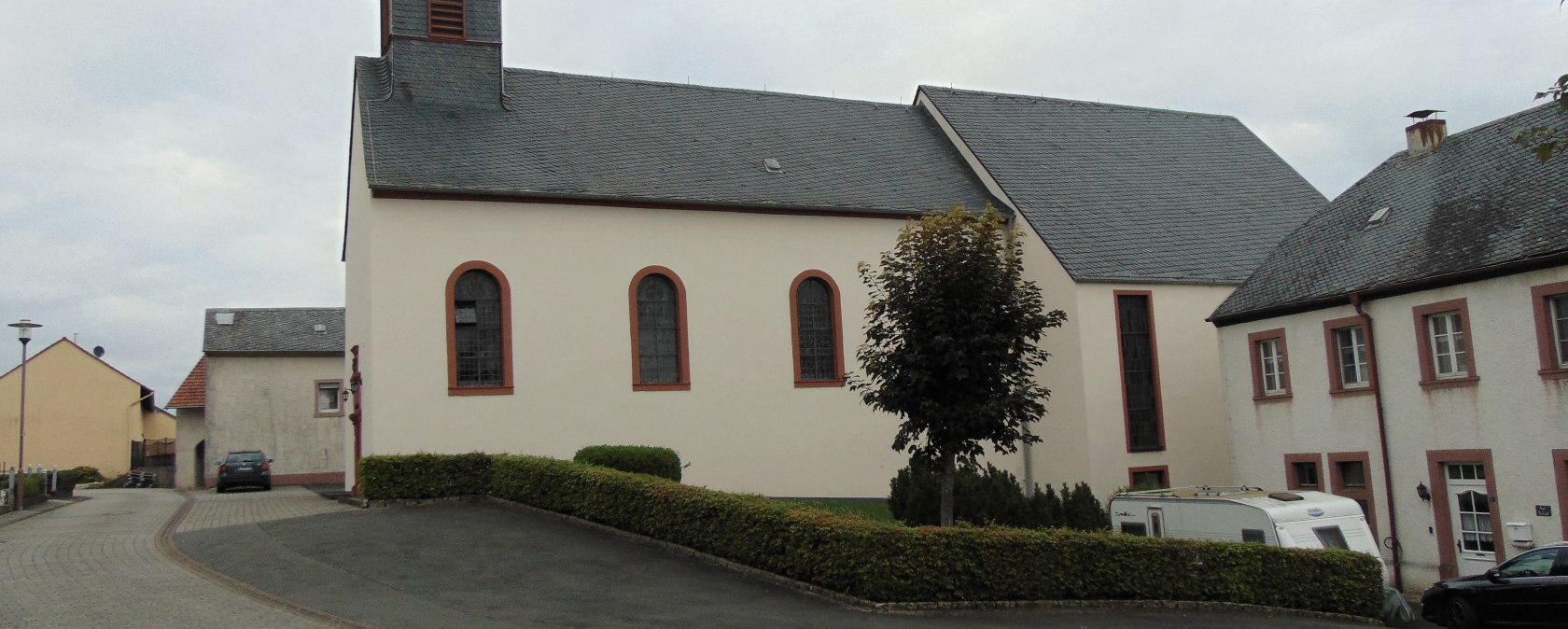 L'&eacute;glise Saint-Urbain &agrave; Gindorf, avec sa tour pointue, entour&eacute;e de maisons d'habitation et d'un chemin pav&eacute;. Un arbre et une caravane sont visibles., &copy; Conny Meier
