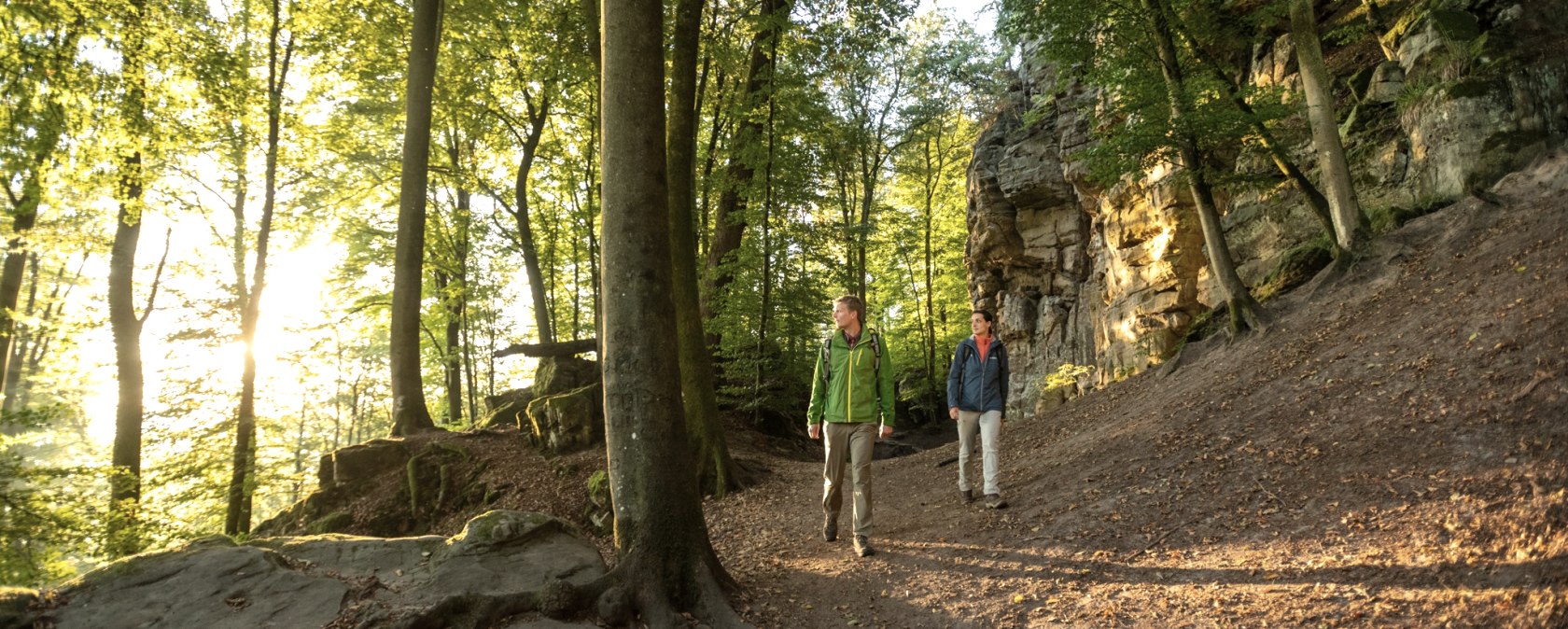 Deux randonneurs marchent sur un chemin forestier le long d'une paroi rocheuse. Le soleil brille &agrave; travers les arbres et baigne la sc&egrave;ne d'une lumi&egrave;re chaude., &copy; Dominik Ketz