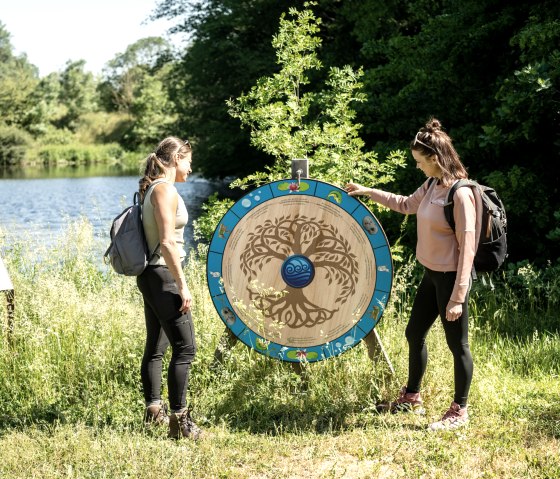 Twee vrouwen met rugzakken kijken naar een decoratief houten wiel aan de oever van een meer, omgeven door groene natuur., © Eifel Tourismus GmbH, Dominik Ketz