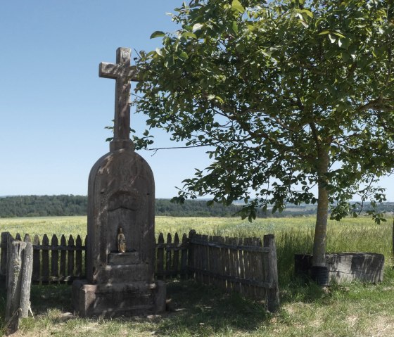 Steinernes Wegekreuz neben einem Baum auf einem Feldweg, umgeben von einem Holzzaun. Im Hintergrund Felder und Wald., &copy; TI Bitburger Land