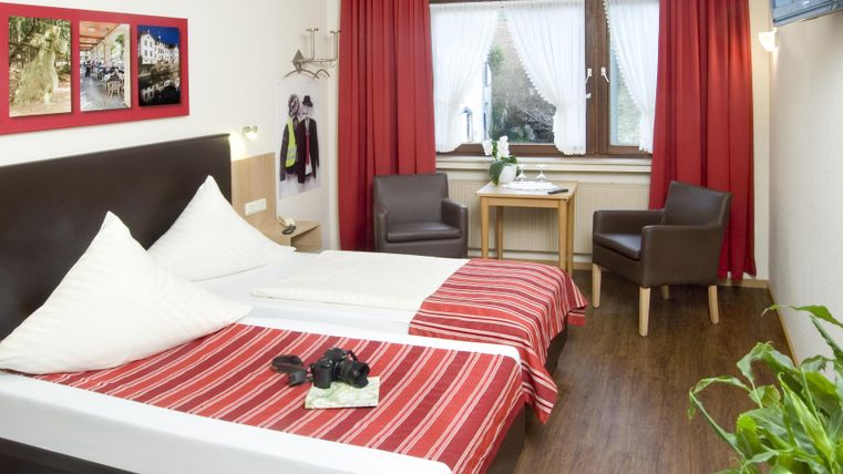 Ein gemütliches Hotelzimmer mit einem Doppelbett in Rot-weiß. Zwei Sessel und ein Tisch ergänzen die Einrichtung, während ein Fenster für Tageslicht sorgt.