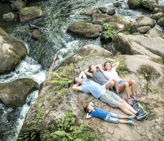 Une famille est allong&eacute;e de mani&egrave;re d&eacute;tendue sur un grand rocher au milieu d'un ruisseau coulant avec des cascades et entour&eacute; d'une verdure luxuriante., &copy; Felsenland S&uuml;deifel Tourismus GmbH