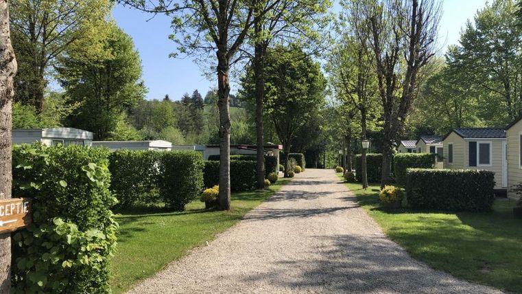 Un chemin calme en gravier, entouré d'arbres et d'arbustes. Des hébergements confortables se trouvent de chaque côté dans un paysage verdoyant.