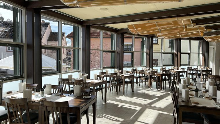 A modern restaurant with many wooden tables and chairs. Large windows let in plenty of light and provide a view of the outside.