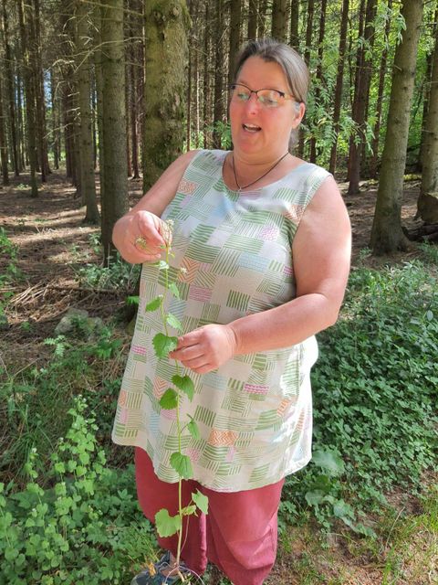 Eine Frau steht im Wald und hält eine Pflanzenranke in der Hand. Sie trägt ein helles Oberteil und hat lange Haare.