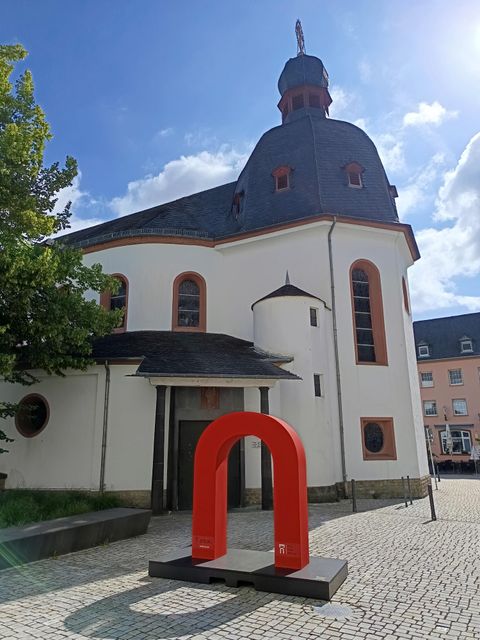 Eine Kirche mit einem modernen roten Kunstwerk im Vordergrund. Der Himmel ist klar und die Umgebung ist hell erleuchtet.