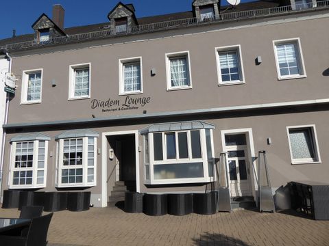 Ein modernes Restaurant namens Diadem Lounge mit großen Fenstern und einem einladenden Eingangsbereich. Die Fassade ist in einem hellen Grauton gehalten und die Umgebung wirkt ordentlich und gepflegt.